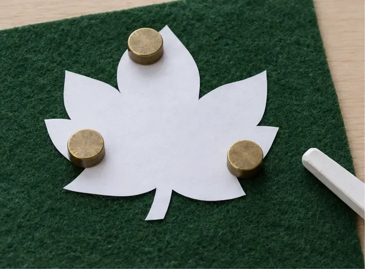 A paper leaf template rests on green felt under three small brass pattern weights, with chalk beside it.