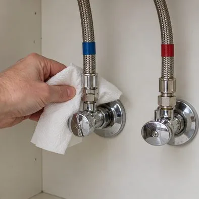 A person checks a shutoff valve and braided faucet supply hose with a dry tissue