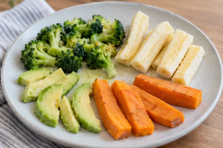 A plate of soft broccoli florets, avocado slices, sweet potato sticks, and soft cheese prepared as finger foods