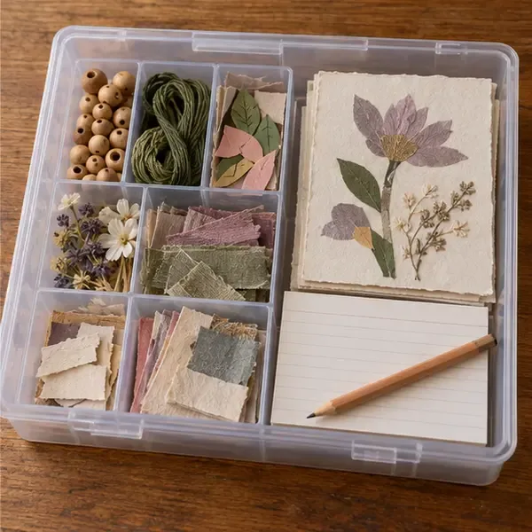 A clear craft box holds sorted beads, thread, paper scraps, an unfinished flower collage, and a blank note card with a pencil.