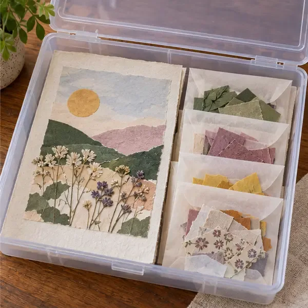 An unfinished botanical paper collage lies flat in a shallow box beside paper scraps sorted into small envelopes.