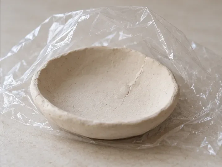 A repaired-looking clay dish rests beneath a loose clear plastic cover on a tabletop.